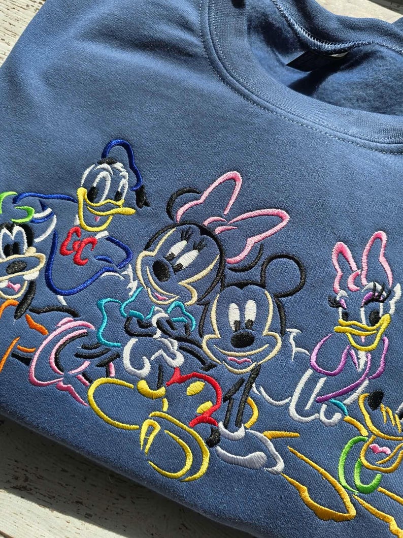 May include: A blue sweatshirt featuring embroidered cartoon characters including Mickey Mouse, Minnie Mouse, Donald Duck, Daisy Duck, and Goofy. The colorful embroidery is set against the solid blue fabric of the crew neck sweatshirt.