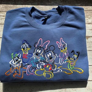 May include: A navy blue sweatshirt featuring an embroidered design of Disney characters, including Mickey and Minnie Mouse, Donald Duck, Daisy Duck, and Pluto. The characters are outlined in various colors.