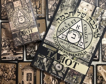May include: A black and white tarot deck with a black box and a guidebook. The guidebook has a black cover with a white triangle and the words "Solve et Coagula" in Hebrew. The cards feature black and white illustrations of tarot symbols.