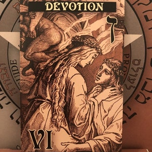 Puede incluir: Una carta del tarot con fondo negro y letras doradas. La carta se titula "Los Amantes" y tiene el n&uacute;mero "VI" en el centro. La imagen de la carta representa a una mujer y un hombre abraz&aacute;ndose, con una figura alada flotando sobre ellos. La carta tambi&eacute;n presenta la palabra "Devoci&oacute;n" en letras doradas y el n&uacute;mero "7" en escritura hebrea.