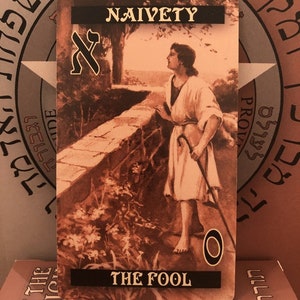 Puede incluir: Una carta del tarot con una imagen en sepia de un hombre de pie en un camino con un bast&oacute;n en la mano. La carta se titula "El Loco" y tiene la palabra "Naivety" en la parte superior. La carta es parte de una baraja de tarot.