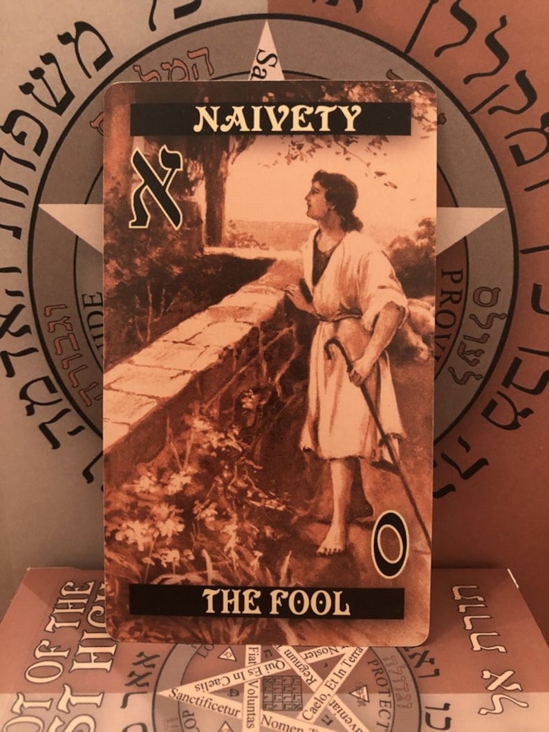 Puede incluir: Una carta del tarot con una imagen en sepia de un hombre de pie en un camino con un bast&oacute;n en la mano. La carta se titula "El Loco" y tiene la palabra "Naivety" en la parte superior. La carta es parte de una baraja de tarot.