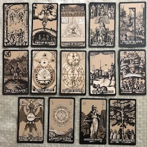 May include: A set of 14 tarot cards featuring black and white illustrations on a beige background. The cards depict various scenes and symbols, including a man with a staff, a woman with a crown, and a tree with a hanging figure. The cards are numbered from 1 to 10 and include the Page, Knight, Queen, and King of Staffs.