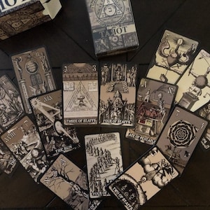 May include: A spread of tarot cards on a dark wood surface. The cards feature black and white illustrations with a gothic aesthetic. The cards include the Three of Staffs, Four of Staffs, Ace of Pentacles, The Sun, Four of Cups, Two of Staffs, and others.