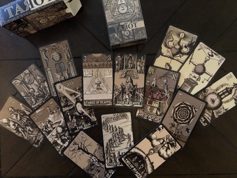 May include: A spread of tarot cards on a dark wood surface. The cards feature black and white illustrations with a gothic aesthetic. The cards include the Three of Staffs, Four of Staffs, Ace of Pentacles, The Sun, Four of Cups, Two of Staffs, and others.