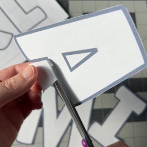 May include: A person is using purple-handled scissors to cut out a white paper shape with a gray border and a gray triangle inside. Other letter shapes are visible in the background on a gray cutting mat.