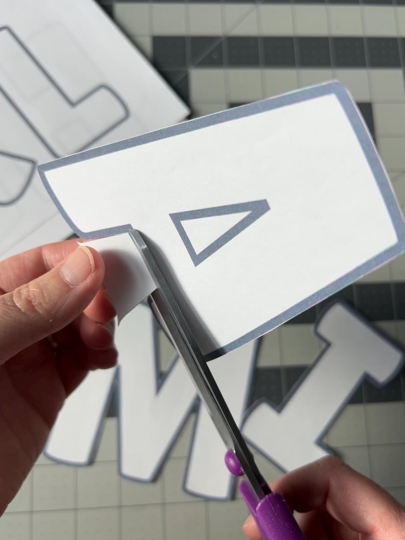 May include: A person is using purple-handled scissors to cut out a white paper shape with a gray border and a gray triangle inside. Other letter shapes are visible in the background on a gray cutting mat.