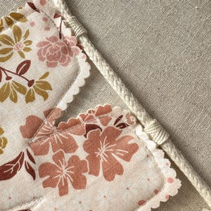 May include: A close-up of a floral banner with a cream-colored braided rope. The banner features a repeating pattern of pink and gold flowers on a cream background. The edges of the banner are scalloped.