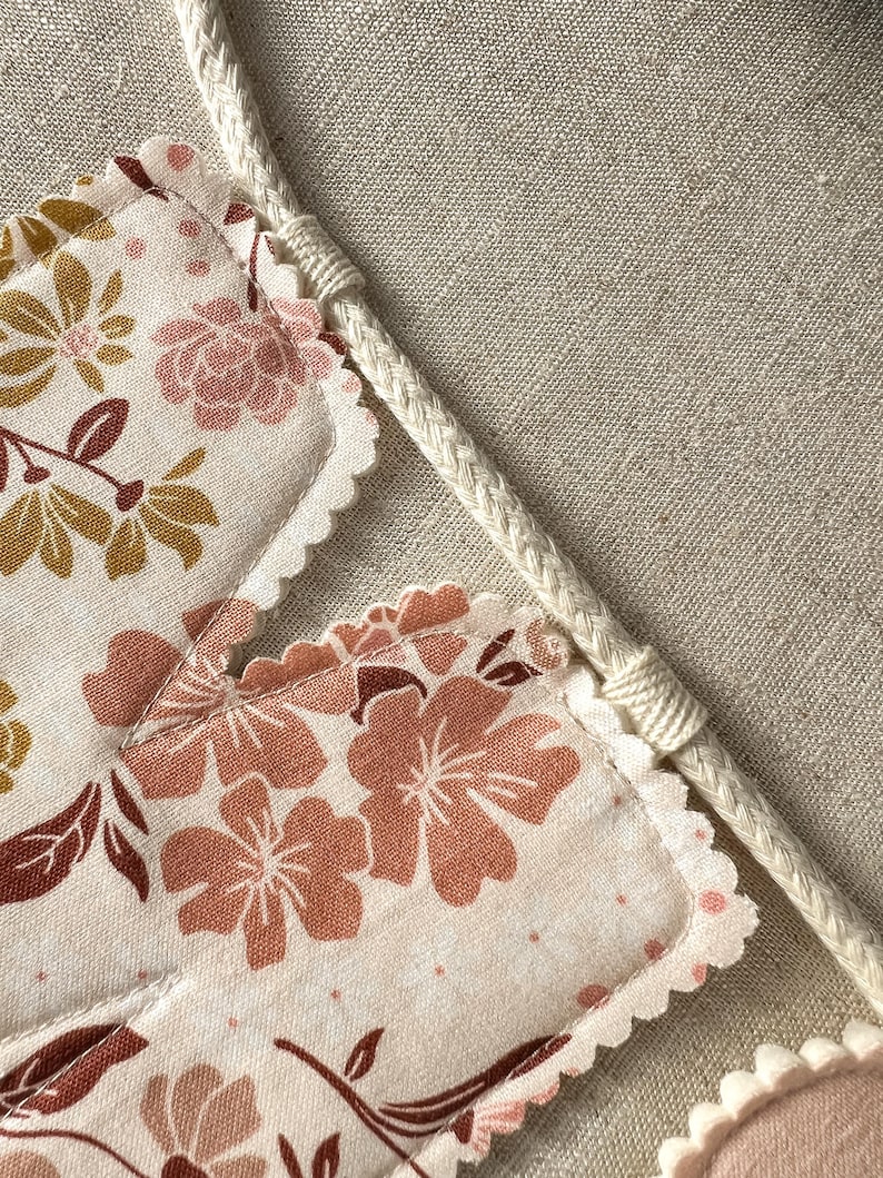 May include: A close-up of a floral banner with a cream-colored braided rope. The banner features a repeating pattern of pink and gold flowers on a cream background. The edges of the banner are scalloped.