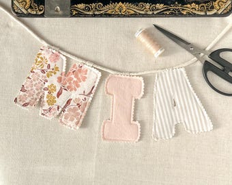May include: A fabric banner spelling the name MIA, featuring letters made from floral patterned fabric, solid light pink fabric, and white and gray striped fabric, all strung on a white cord with a spool of thread and black scissors.
