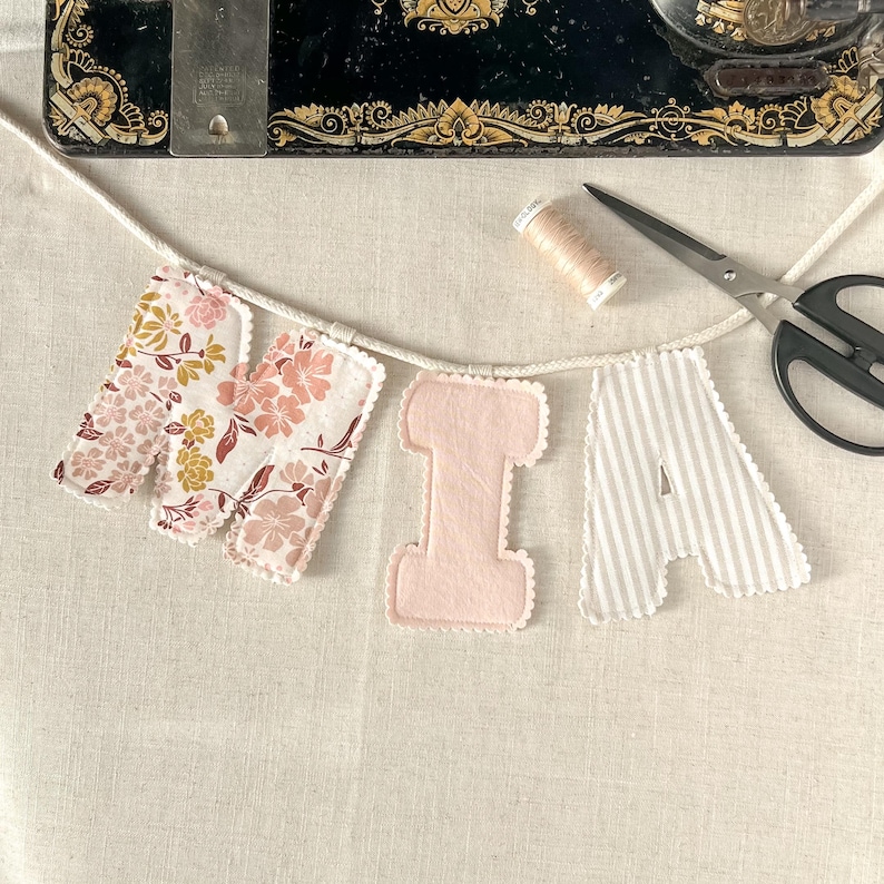 May include: A handmade banner spelling "MIA" with fabric letters. The "M" features floral patterns, the "I" is solid pink, and the "A" has gray and white stripes. A spool of thread and scissors are also visible.