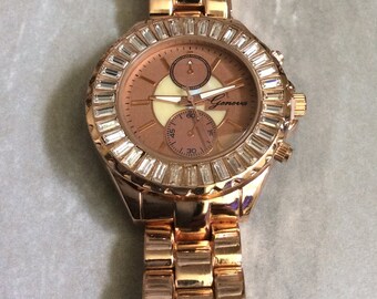 May include: Rose gold-toned Geneva watch with a crystal-studded bezel. The watch features a rose-colored face with sub-dials and the brand name "Geneva". The band is made of matching rose gold links.