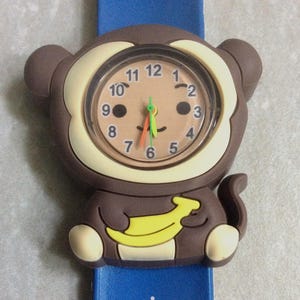 Könnte beinhalten: Eine Armbanduhr mit einem braunen, affenförmigen Zifferblatt, das eine gelbe Banane hält. Das Zifferblatt zeigt schwarze Zahlen von 1 bis 12 sowie orangefarbene und grüne Zeiger. Das blaue Armband ist mit kleinen hellblauen Pfotenabdrücken verziert.