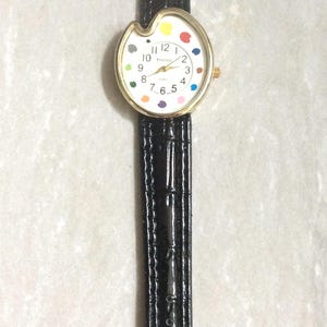 May include: A wristwatch with a black leather band and a gold-colored case. The watch face is white with colorful dots marking the hours. The watch has a unique, artistic design.