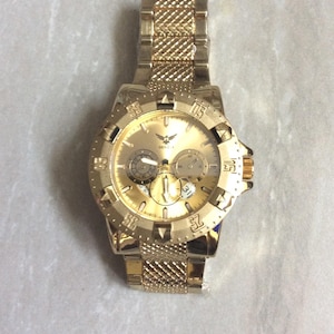 May include: Gold-coloured Geneva watch with a textured strap and face. The watch features a round dial with multiple sub-dials and a textured bezel. The watch has a gold-coloured eagle logo at the top of the face.