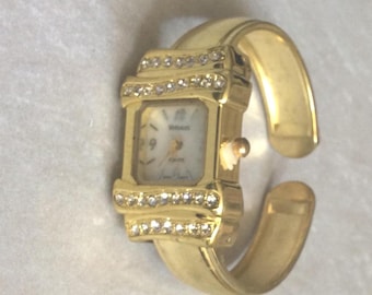 Può includere: Un orologio ad anello dorato con quadrante rettangolare. Il quadrante è bianco con numeri e lancette nere. L'orologio è impreziosito da piccoli cristalli trasparenti. Il cinturino ha un design ampio e aperto.