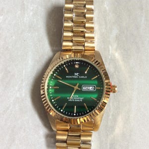 May include: Gold-toned wristwatch with a green dial. The watch features a gold band and a round face with the text "MONTRES CARLO". The dial displays the day and date. The watch has a textured bezel and a small diamond accent.
