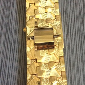 May include: A rectangular, gold-coloured lighter with a textured surface. The lighter features a geometric pattern of various shapes and textures, including squares, triangles, and lines. The lighter is closed, with a visible hinge and clasp.