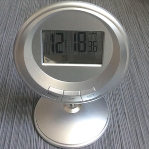 Puede incluir: Un reloj digital plateado que muestra la hora 12:18:36. El reloj tiene una esfera redonda y una base circular. Cuenta con tres botones etiquetados como "MODE", "UP" y "SET".