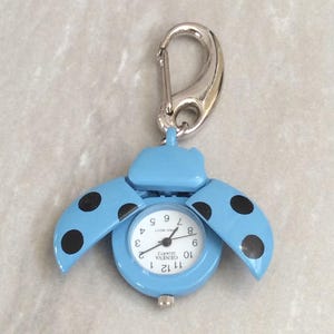 May include: A light blue ladybug-shaped pocket watch with black polka dots on the wings. The watch face is white with black numerals and hands. It has a silver-colored carabiner clip for attachment.