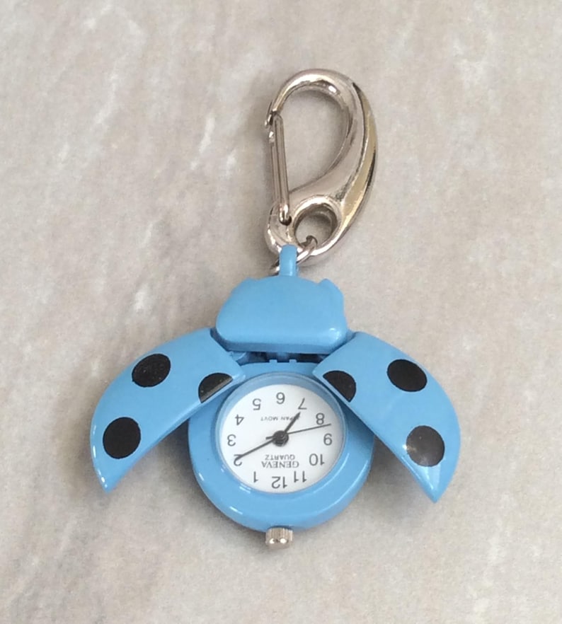 May include: A light blue ladybug-shaped pocket watch with black polka dots on the wings. The watch face is white with black numerals and hands. It has a silver-colored carabiner clip for attachment.