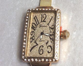 Può includere: Orologio Geneva rettangolare dorato al quarzo con quadrante glitterato, numeri neri e lunetta bordata di piccoli cristalli. L'orologio ha un cinturino in maglia dorata e una corona strutturata. Il quadrante mostra l'ora.