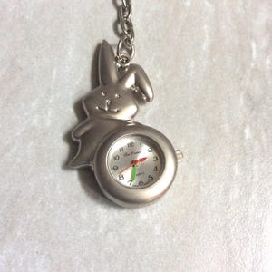 Könnte beinhalten: Eine silberfarbene Taschenuhr mit einem hasenförmigen Rahmen. Das Zifferblatt hat schwarze Ziffern, einen roten Sekundenzeiger und einen grünen Minutenzeiger. Die Uhr ist an einer silbernen Kette befestigt.