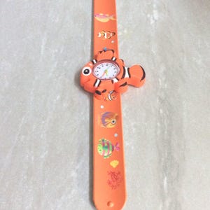 May include: Orange children's watch with a fish design. The watch face is white with black numbers and hands. The band is orange and decorated with colorful fish illustrations. The watch is designed to be worn on the wrist.