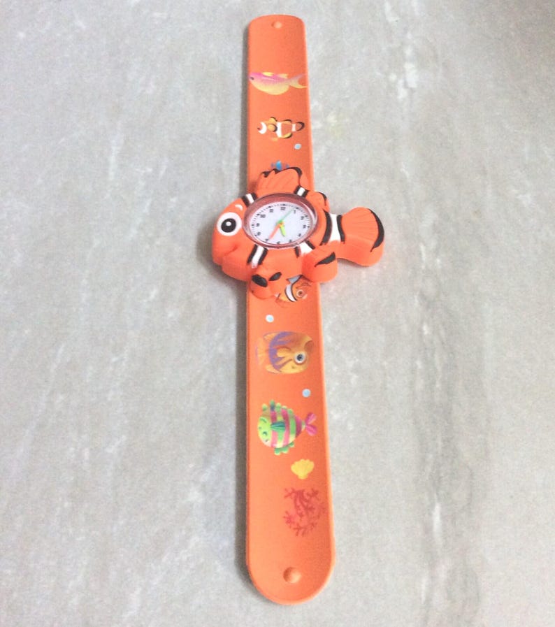 May include: Orange children's watch with a fish design. The watch face is white with black numbers and hands. The band is orange and decorated with colorful fish illustrations. The watch is designed to be worn on the wrist.
