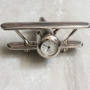 May include: A silver-toned metal biplane desk clock with a polished finish. The front of the airplane features a round analog clock face with a white background, black numerals, and the word Geneva printed in black above the center hands.