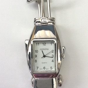 May include: A silver-toned quartz watch shaped like a golf bag with clubs protruding from the top. The rectangular white watch face features black numerals from 1 to 12, the text GENEVA, QUARTZ, and JAPAN MOVT. The metallic casing has a polished finish.