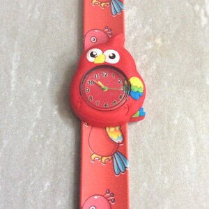 Peut inclure: Une montre rouge pour enfants avec un visage en forme d'oiseau et un bracelet rouge assorti. Le cadran de la montre affiche l'heure, et le bracelet présente des illustrations d'oiseaux répétées de différentes couleurs. La montre est conçue pour les enfants.