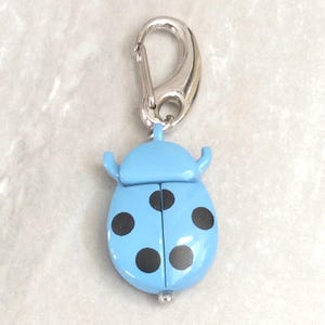May include: A light blue ladybug-shaped keychain with black spots and a silver-colored clasp. The ladybug has two small antennae and a hinged body. The keychain is a decorative accessory.