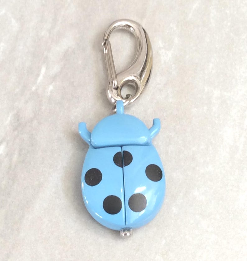 May include: A light blue ladybug-shaped keychain with black spots and a silver-colored clasp. The ladybug has two small antennae and a hinged body. The keychain is a decorative accessory.
