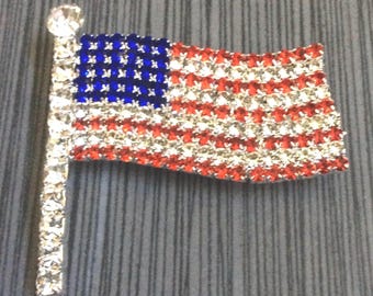 May include: A brooch shaped like the American flag, crafted from sparkling crystals. The flag design includes red and clear crystal stripes, a blue crystal field with white stars, and a crystal-covered pole. A patriotic accessory.