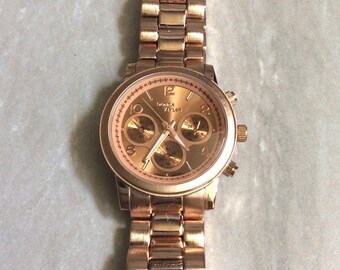 May include: Rose gold-toned wristwatch with a matching band. The watch face features three sub-dials and a date display. The watch has a round face with a rose gold bezel and a link-style band.