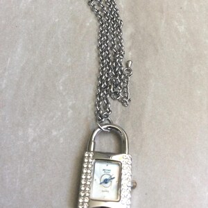 Könnte beinhalten: Eine silberfarbene, schlossförmige Anhängeruhr mit passender Kette. Das Zifferblatt der Uhr ist weiß mit blauen Zeigern und einem kleinen Zifferblatt. Die Uhr ist mit kleinen, klaren Kristallen um das Zifferblatt herum besetzt.