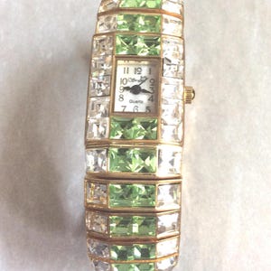Puede incluir: Un reloj de cuarzo rectangular con una correa de metal dorado y una esfera blanca. La correa está adornada con filas alternas de cristales transparentes y verdes. La esfera muestra la hora y la palabra "Quartz".