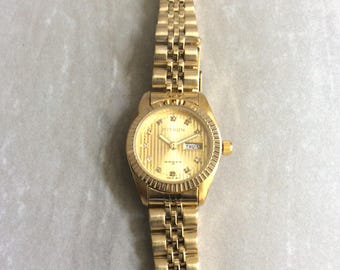 May include: A gold-toned wristwatch with a matching band. The watch face features a textured design, diamond accents, and the day and date display. The watch is labeled "Mithon".