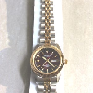 Puede incluir: Un reloj de pulsera Swanson Japan con una correa plateada y dorada y una esfera burdeos. El reloj tiene un bisel y agujas doradas, con un pequeño diamante a las 12 en punto. El día de la semana se muestra a las 3 en punto.