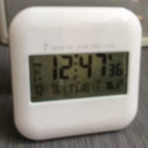 Pode incluir: Um relógio digital branco e quadrado que mostra a hora 12:47:36. O relógio também mostra a data, o dia e a temperatura. O texto "RADIO CONTROLLED" está impresso na parte superior do relógio.