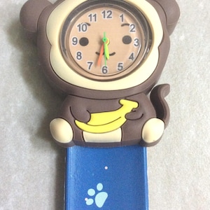 Op de afbeelding: Een kinderhorloge met een bruin apenontwerp met een gele banaan. De wijzerplaat heeft zwarte cijfers en een lachend gezicht. De blauwe band heeft pootafdrukken.