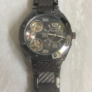 Op de afbeelding: Een donkergrijze Elgin-horloge met een zwarte band. De wijzerplaat heeft een donkergrijze achtergrond met zilveren wijzers en cijfers. Het horloge heeft drie sub-wijzerplaten en een getextureerde band.