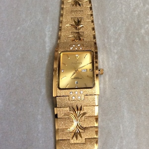 Può includere: Orologio da polso color oro con quadrante rettangolare e cinturino strutturato. Il quadrante mostra il giorno e la data ed è adornato da piccoli cristalli. Il cinturino presenta motivi decorativi floreali.