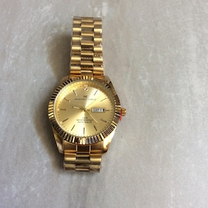 May include: A gold-toned wristwatch with a matching band. The watch face displays the day and date, with the text "MONTRES CARLO" and "SAT 1". The watch has a textured bezel and a red accent on the crown.