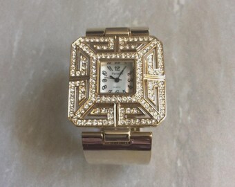 Può includere: Un orologio da polso quadrato color oro con quadrante bianco, numeri e lancette neri. La lunetta è decorata con cristalli trasparenti disposti in un motivo geometrico a greca. L'orologio presenta un ampio cinturino in metallo dorato lucido.