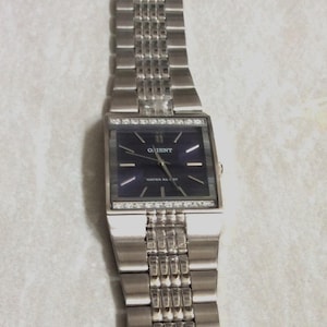May include: A rectangular silver-tone wristwatch featuring a dark blue dial with the Orient logo and silver hour markers. The watch case is accented with a row of clear stones at the top and bottom, attached to a multi-link silver metal bracelet.
