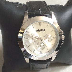 Könnte beinhalten: Silberfarbene Unlisted-Uhr mit hellem Zifferblatt und schwarzem Lederarmband. Die Uhr verfügt über drei Hilfszifferblätter und eine Datumsanzeige. Die Uhr wird auf einem schwarzen Kissen präsentiert.