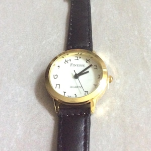 May include: A gold-coloured wristwatch with a white face and black hands. The watch features Hebrew numerals and the word "Finesse" above the centre. The watch has a black leather strap.
