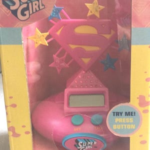 May include: Pink Supergirl LCD alarm clock in a box. The clock features a pink Superman logo, yellow stars, and blue stars. The clock has a digital display and buttons labeled "SET" and "TALY". The box has the text "TRY ME! PRESS BUTTON".